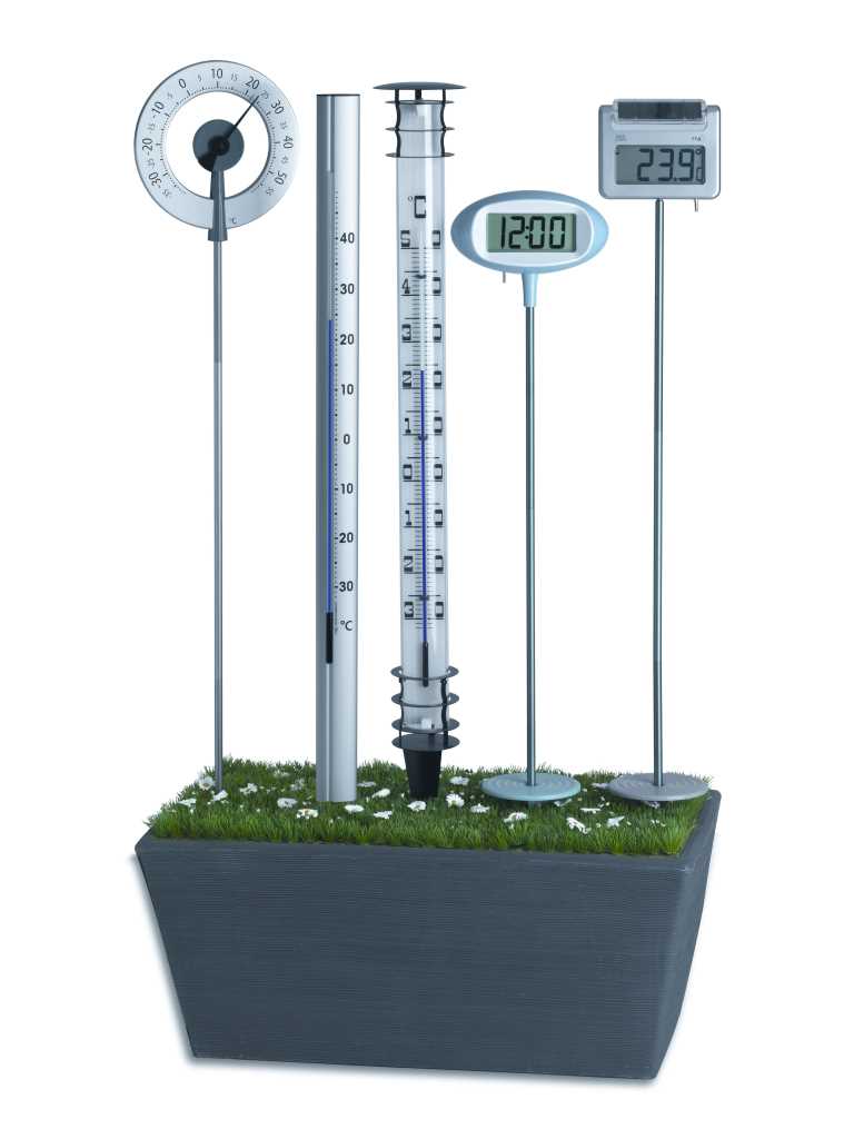 Dunkelgrauer Pflanzkasten mit Gras und weißen Blüten, darin vier Thermometer und eine Digitaluhr auf Stäben